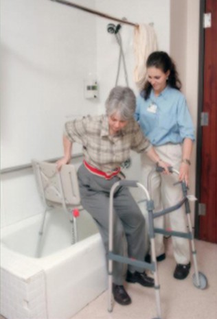 A caregiver assists an elderly woman using a walker to safely enter a bathtub.