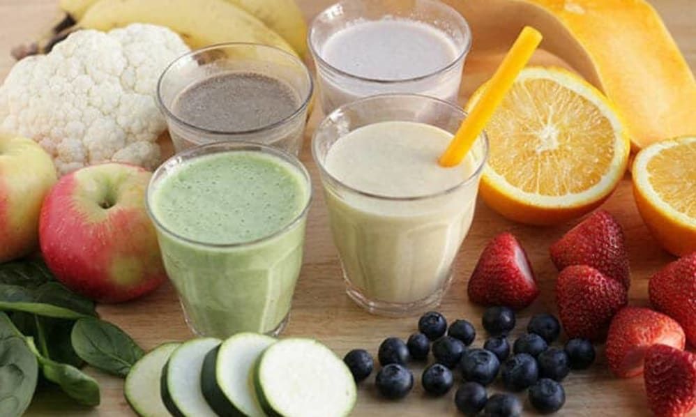 A variety of fruit smoothies surrounded by fresh ingredients like bananas, blueberries, orange, strawberries, and cucumber slices.