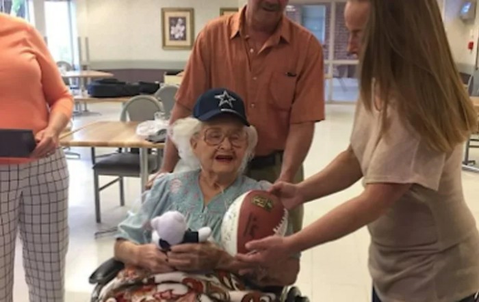 An elderly woman in a wheelchair smiles as she holds a football, surrounded by two people indoors.