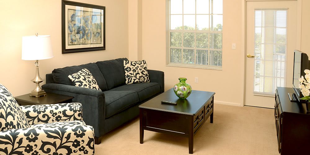 A cozy living room with a dark sofa and patterned cushions, featuring a wooden coffee table with a decorative vase, next to a large window.