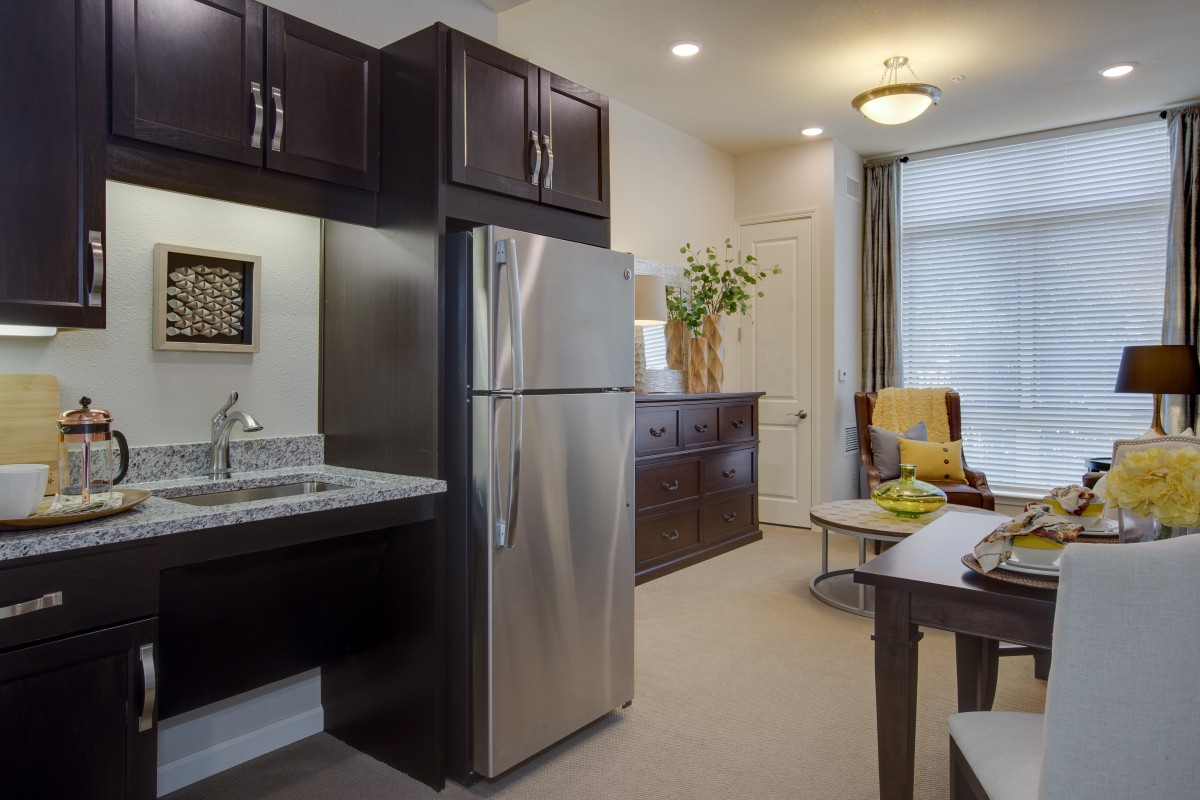 A modern kitchen and living area in an apartment, featuring a stainless steel refrigerator, dark wood cabinets, and comfortable seating.