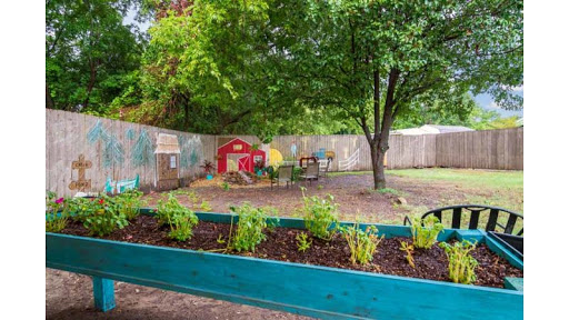 A vibrant outdoor garden area featuring raised beds with plants, a colorful play structure, and a large tree providing shade.