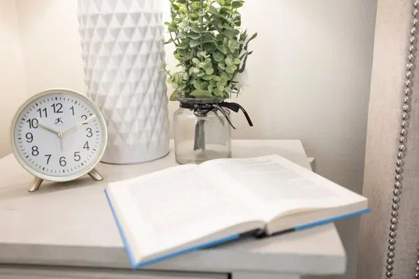 A cozy bedside setup with an open book, a clock, a lamp, and a plant on the table.