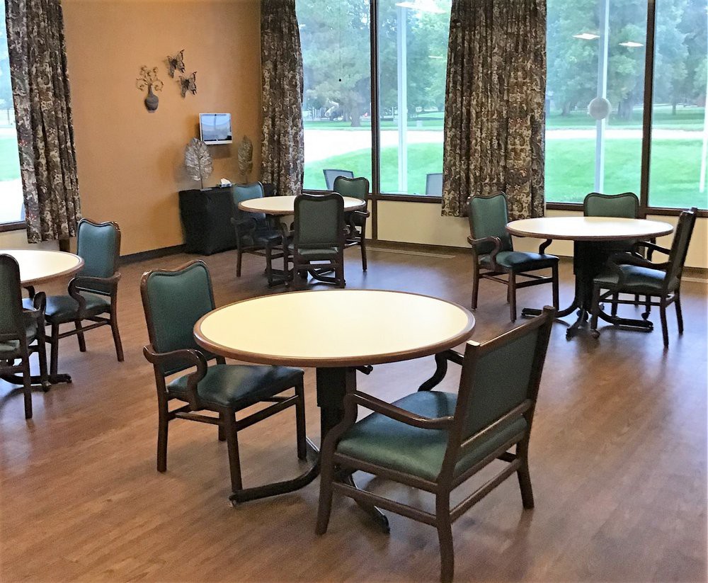 A dining area with multiple round tables and chairs, large windows, and curtains.