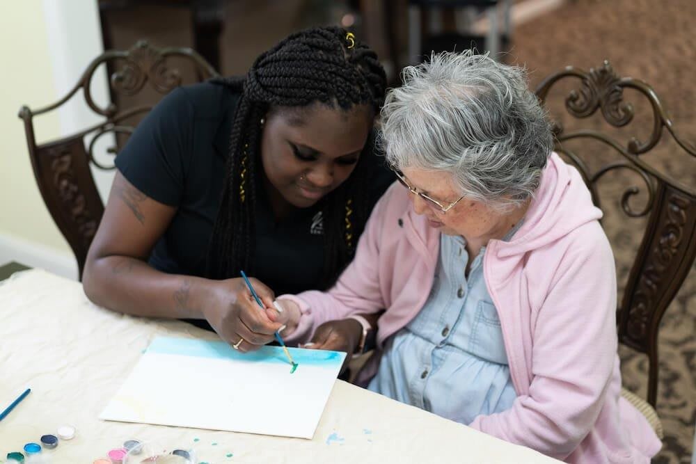 A caregiver assists an elderly woman with a painting project.