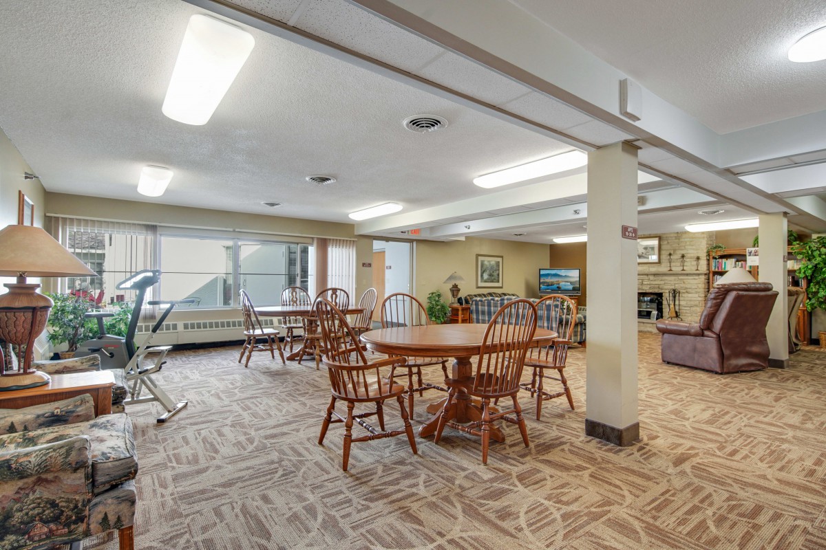 A spacious room with wooden tables and chairs, carpeted flooring, and exercise equipment in the corner.