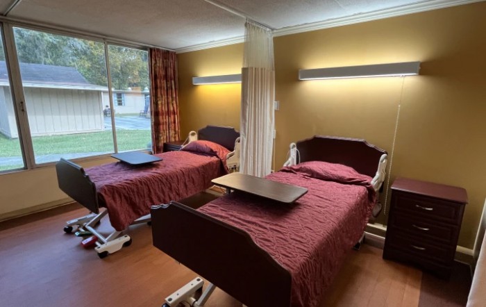 A hospital room with two beds separated by a curtain, featuring large windows and tray tables.