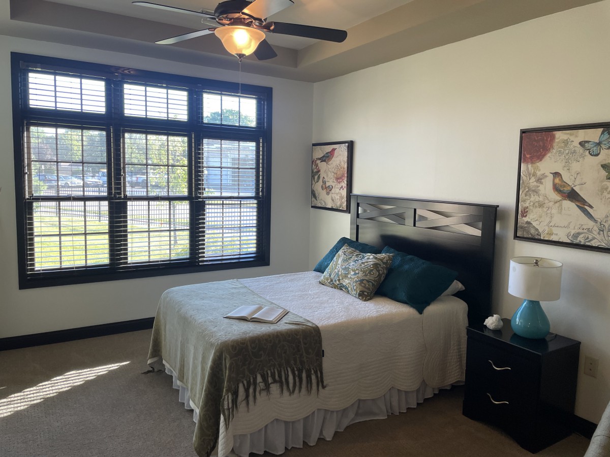 A cozy bedroom with a large window and ceiling fan, featuring a neatly made bed with decorative pillows.