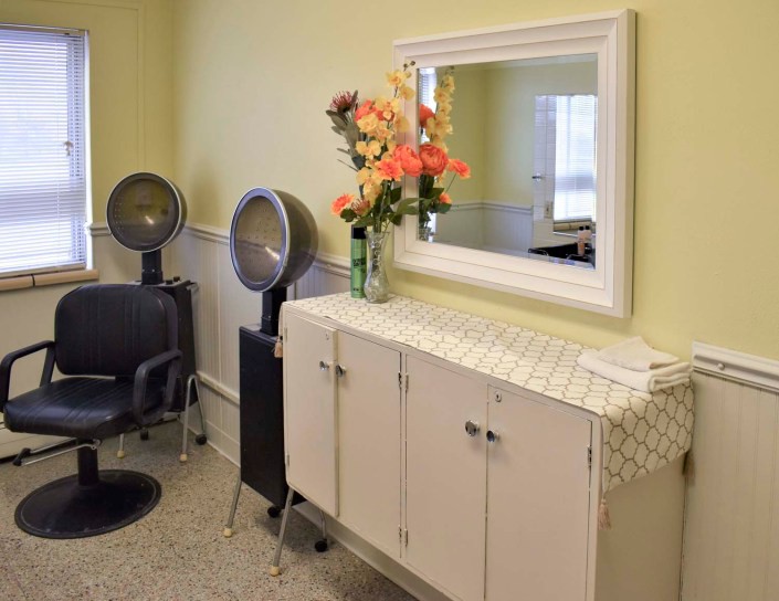 A well-organized salon corner with chairs, hairdryers, a mirror, and fresh flowers on the cabinet.