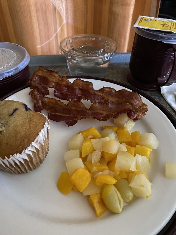 A hospital breakfast tray featuring crispy bacon, a muffin, mixed fruit salad, and juice.
