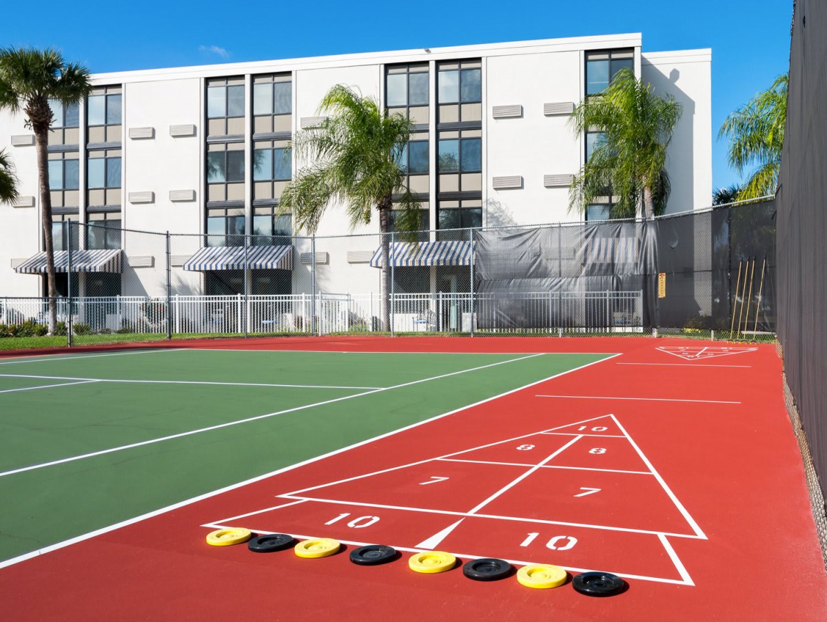 An outdoor recreational area featuring a tennis court and shuffleboard setup, with a modern building and palm trees in the background.