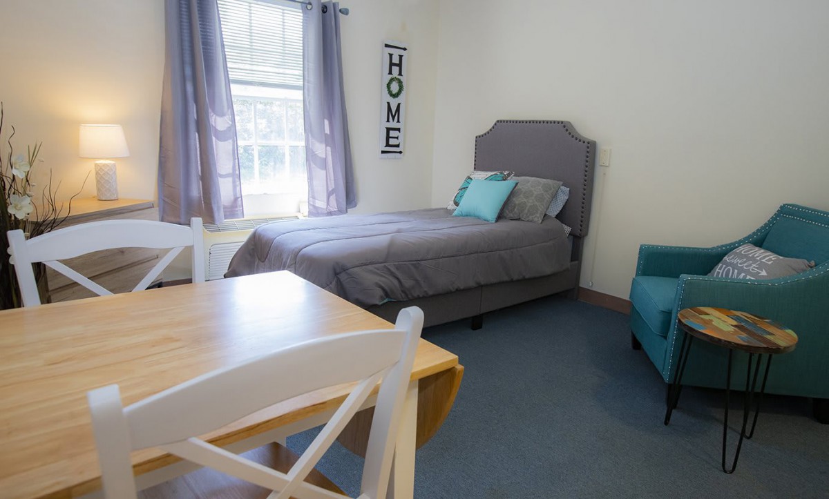 A cozy bedroom interior featuring a neatly made bed with gray bedding, a wooden table and chair set, and a bright window with curtains.