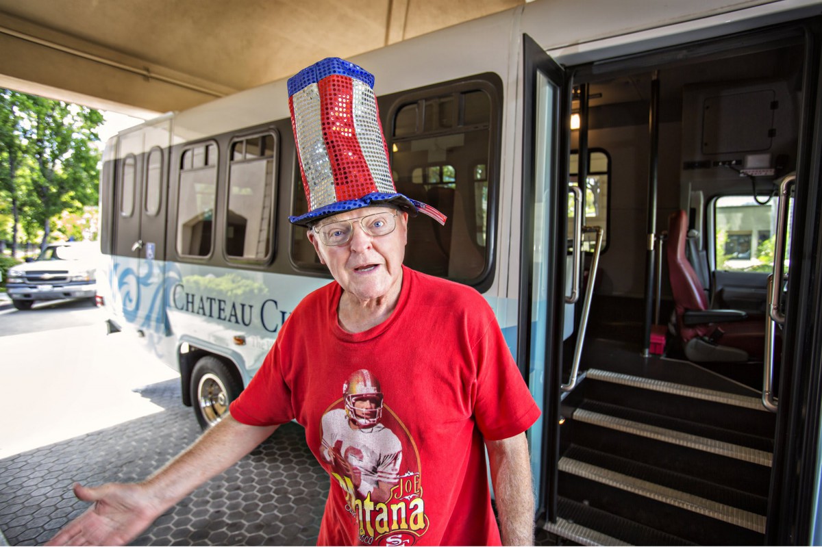 An elderly person wearing a patriotic hat and t-shirt stands in front of a bus, smiling.