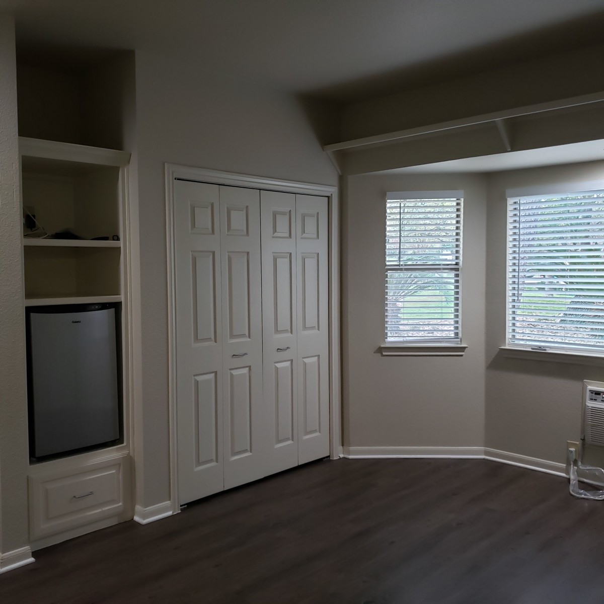 An empty room with built-in shelves and a closet, featuring large windows and wood flooring.