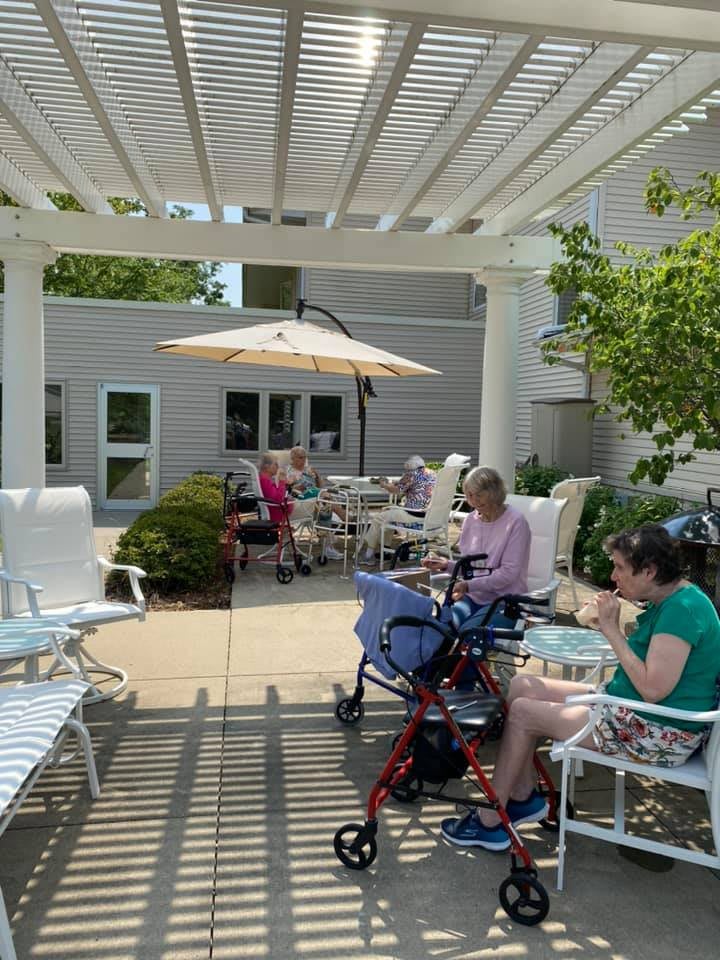A sunny outdoor patio with people sitting, some using wheelchairs and walkers, enjoying a pleasant day.