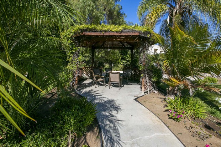 A tranquil garden scene featuring a pergola with outdoor seating, surrounded by lush greenery and tropical plants.