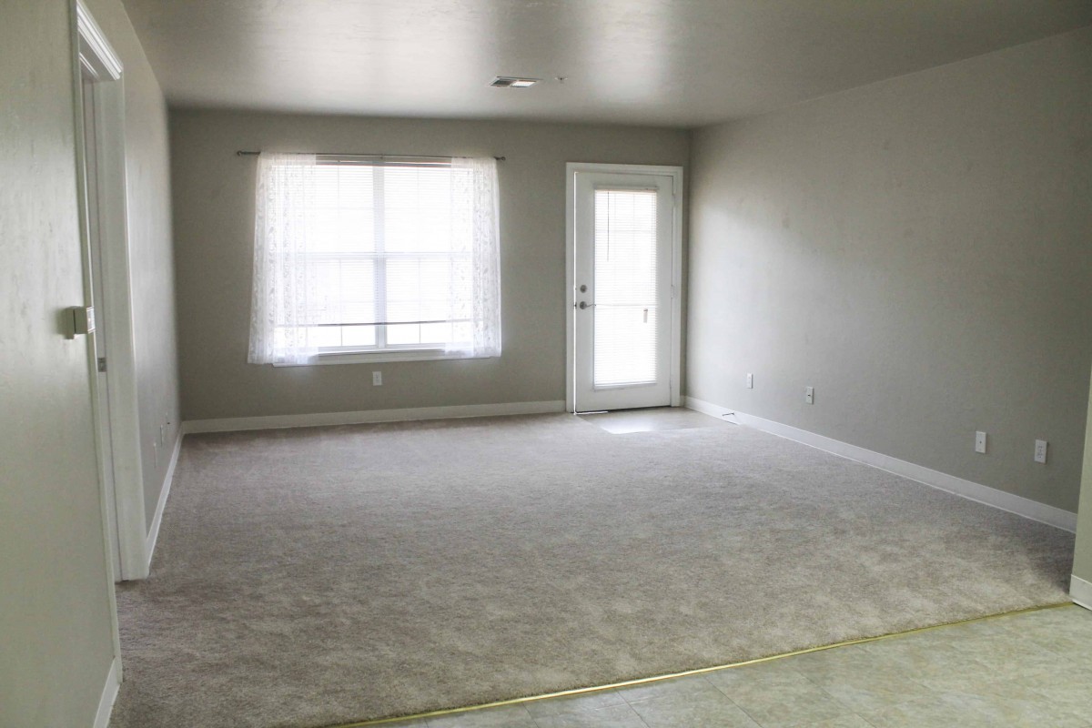 An empty, carpeted living room with a large window and a glass door.