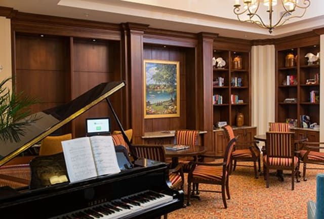 A sophisticated library interior featuring a grand piano with sheet music, surrounded by bookshelves and a table with chairs.