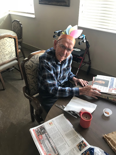 An elderly man enjoys reading a newspaper while wearing a colorful hat indoors.