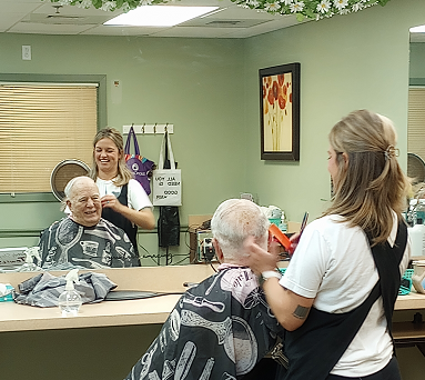 A hairstylist gives a haircut to an elderly client while their reflection is visible in the mirror.