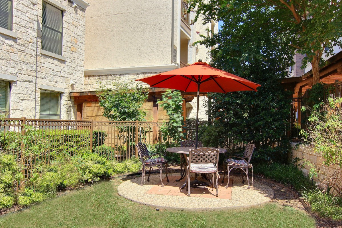 A cozy outdoor patio area with a red umbrella, table, and chairs, surrounded by greenery.