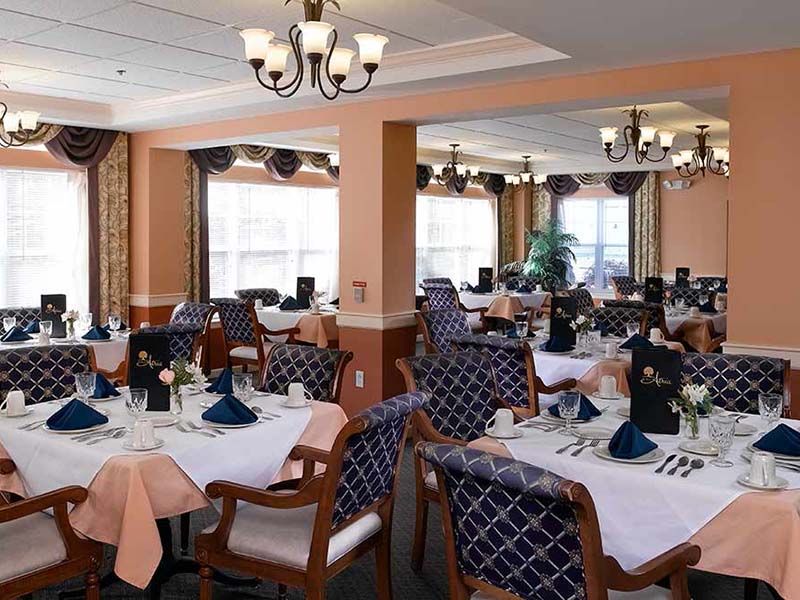 An elegant dining room with neatly arranged tables and chairs, featuring chandeliers and natural lighting.