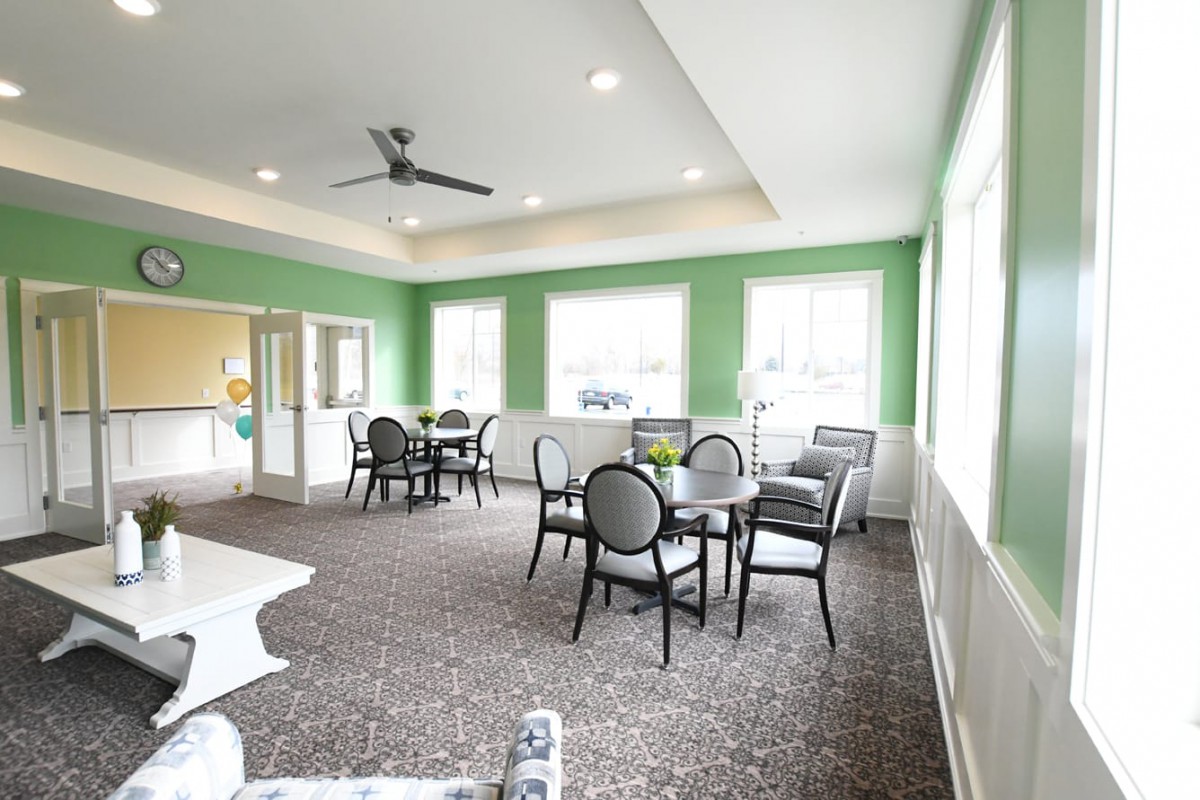 A brightly lit dining area with green walls, featuring several tables and chairs, carpeted flooring, and large windows.