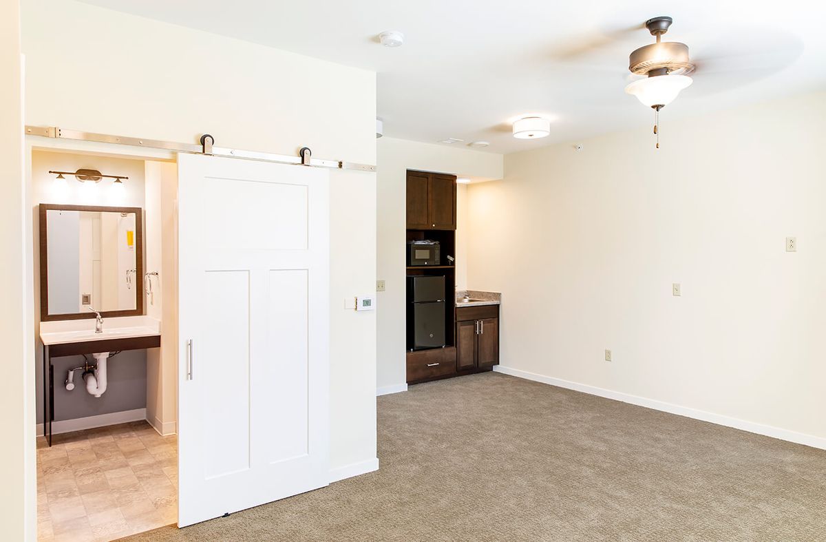 A minimalistic interior room with a ceiling fan and carpet flooring, featuring a sliding door and adjacent kitchenette.