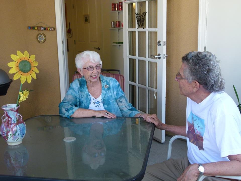 An elderly woman and a man having a conversation at a glass table on a patio.