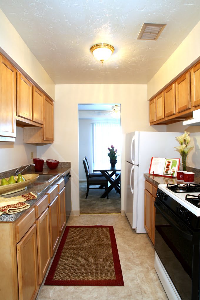 A modern apartment kitchen with wooden cabinets and white appliances, leading to a dining area.
