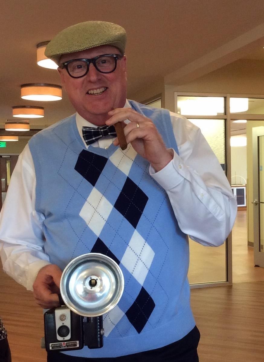 A man wearing a hat, glasses, and a bow tie holds a cigar and stands indoors.
