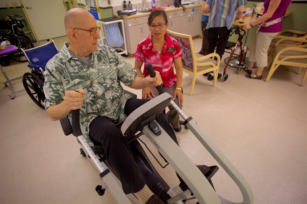 A person receives guidance while using a recumbent exercise machine in a healthcare setting.