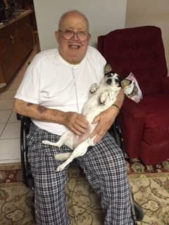 A man sitting in a wheelchair, smiling and holding a small dog indoors.