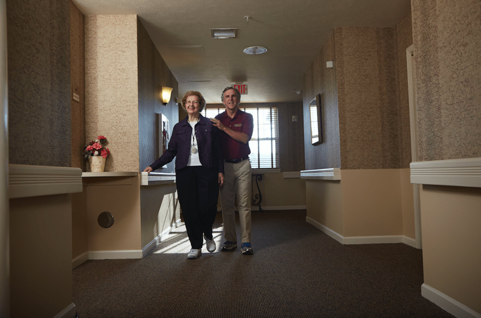 An elderly woman receiving assistance while walking down a hallway.