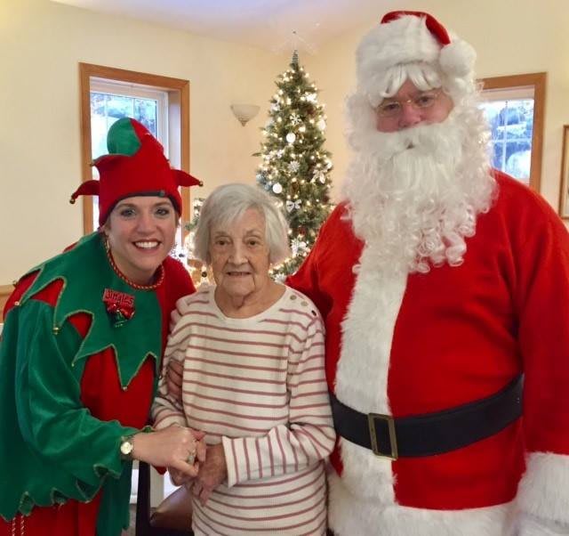 An elderly woman is joyfully posing with Santa and an elf during a Christmas celebration.