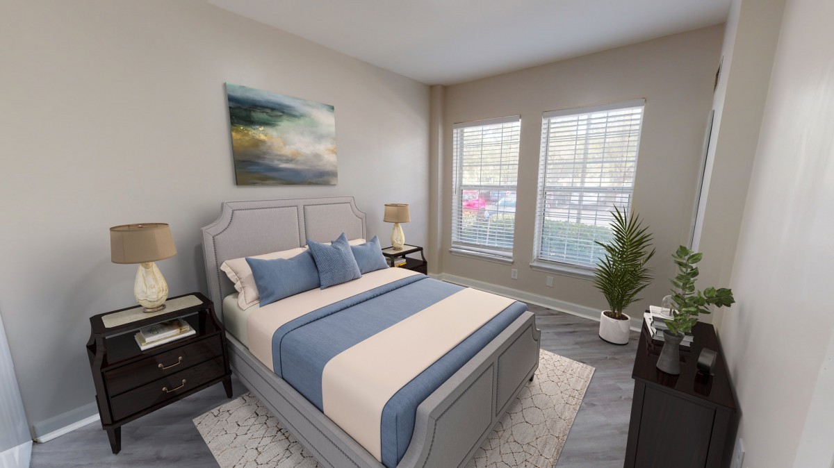 A modern bedroom with a neatly made bed, featuring blue and beige bedding, artwork on the wall, large windows allowing natural light, and a potted plant in the corner.