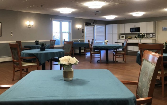 A communal dining area with tables and chairs, featuring a kitchen and decorative flowers on the tables.