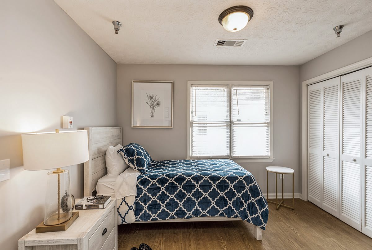 A cozy bedroom with a neatly made bed featuring a blue geometric-patterned duvet, flanked by a nightstand with a lamp, and artwork hanging on the wall. The room has ample natural light coming through the window.