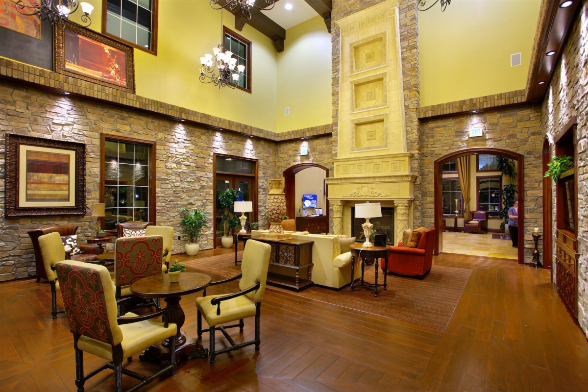 A warm and inviting living room featuring a tall stone fireplace, wooden floors, and elegant furnishings under a high ceiling with a chandelier.
