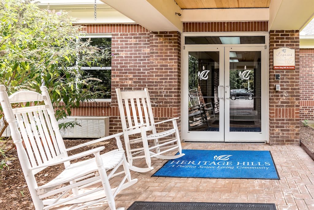 An inviting entrance with white rocking chairs and a welcome mat outside a brick building.