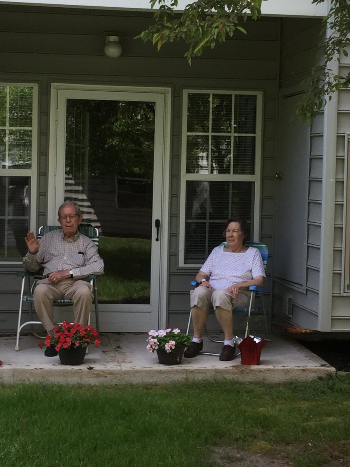 A couple sitting on chairs outside their house, surrounded by potted flowers.