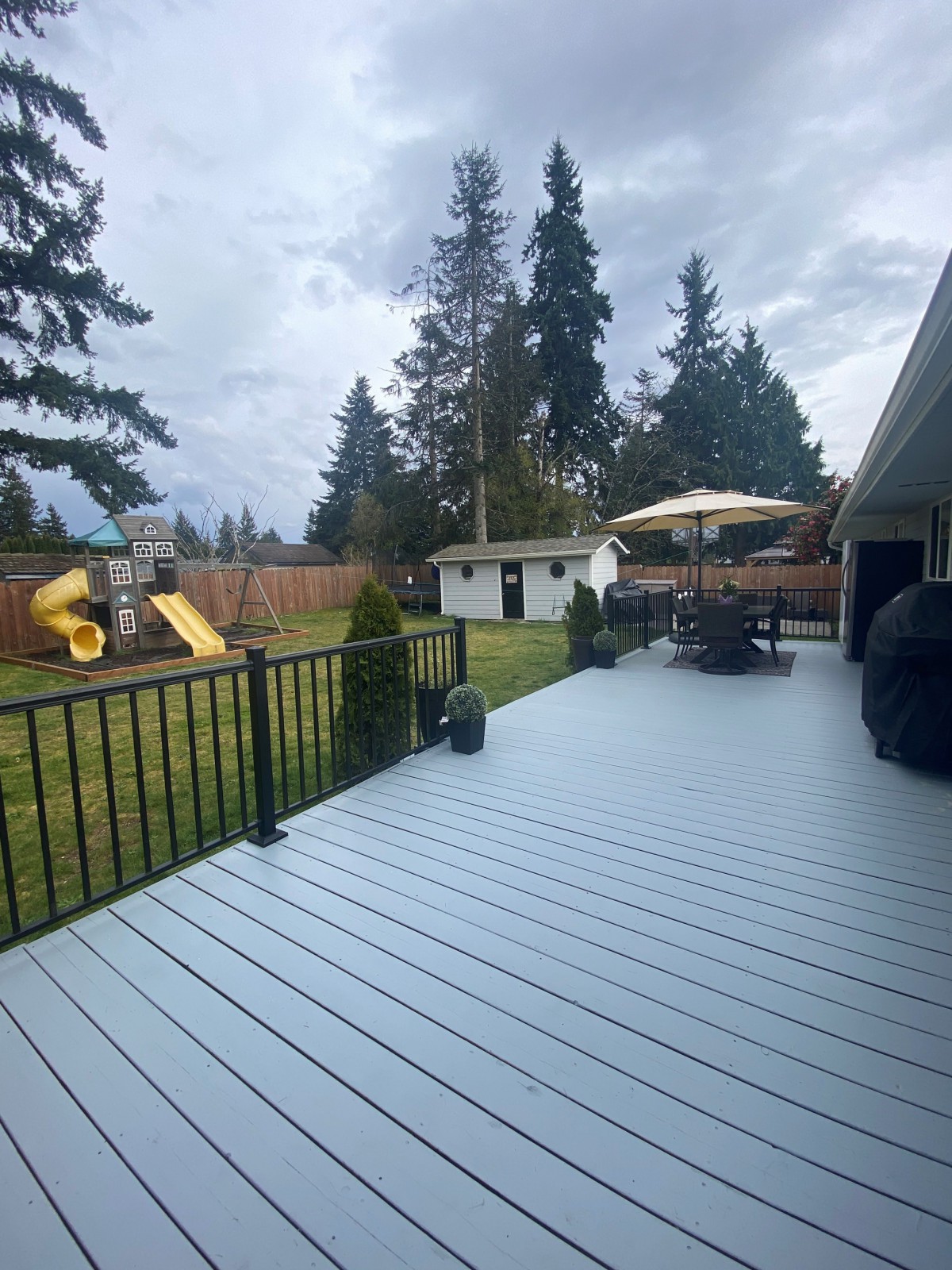 A spacious backyard featuring a wooden deck with outdoor furniture and an umbrella, alongside a children's playground set on a cloudy day.