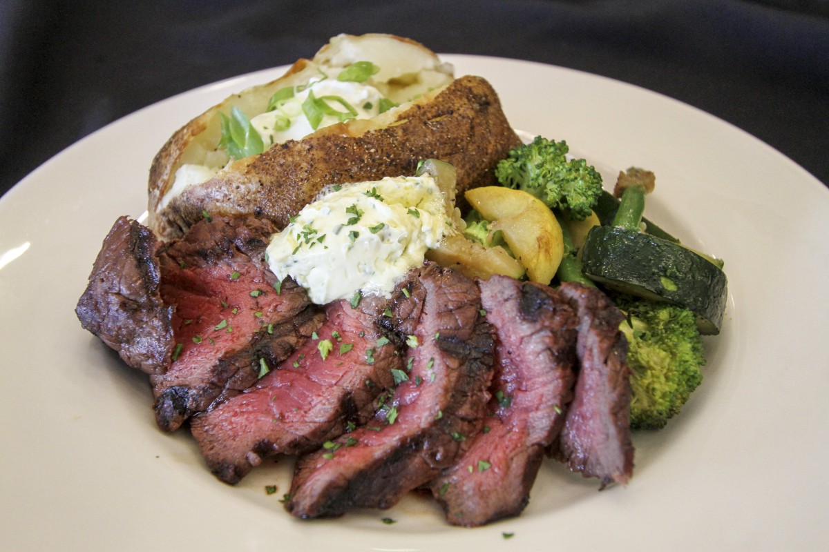 A plate featuring sliced steak, a baked potato topped with sour cream, and steamed vegetables including broccoli and zucchini.
