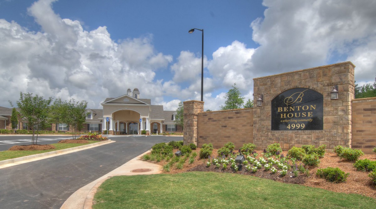 A welcoming entrance to a large building with well-maintained landscaping and a sign displaying its name under a partly cloudy sky.