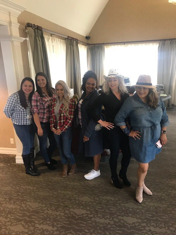 A group of women posing together indoors, wearing casual outfits and a couple of them wearing hats.