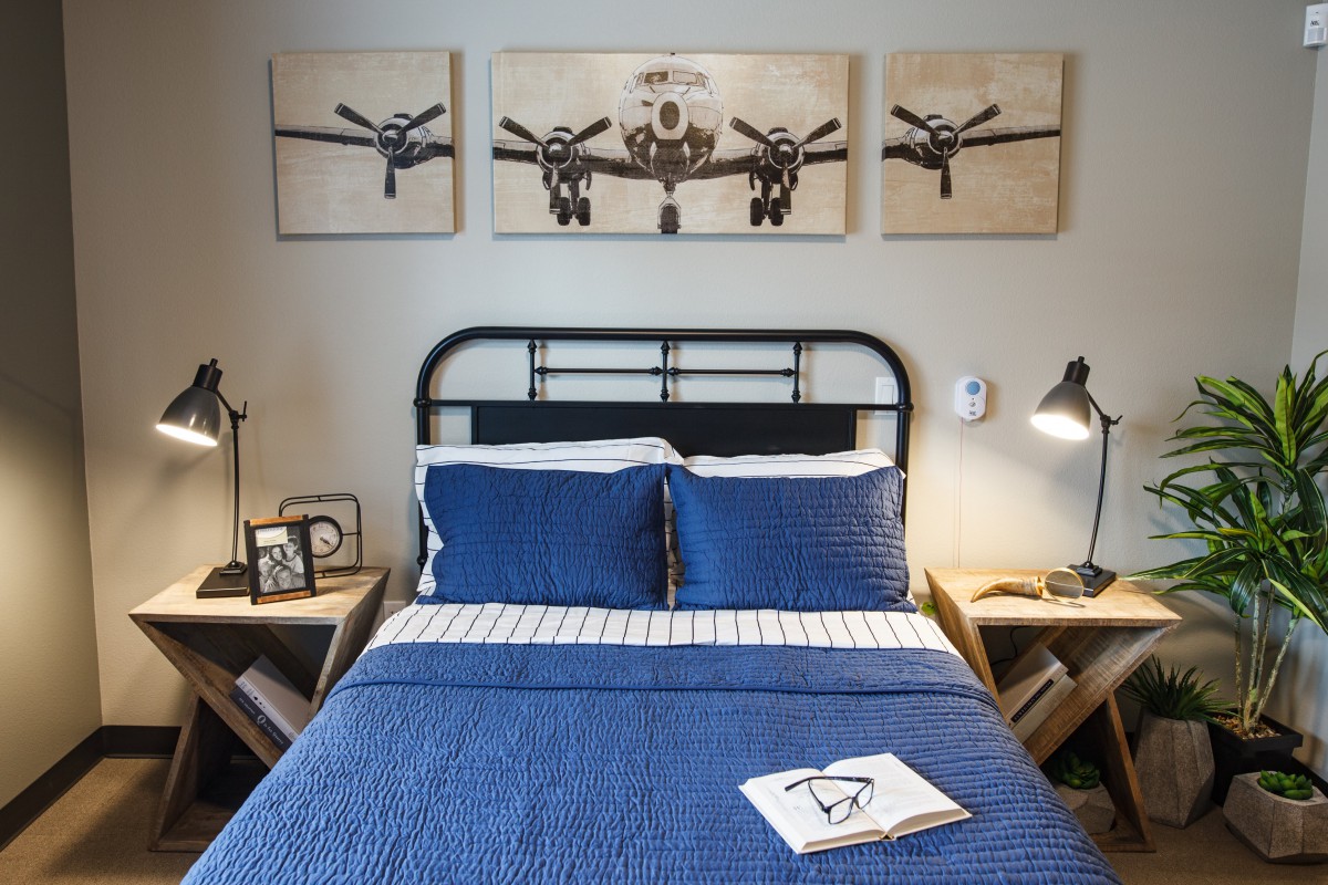 A stylish bedroom features a black metal bedframe with blue bedding, flanked by wooden nightstands. Above the bed, a triptych of aeroplane artwork adds an industrial touch.