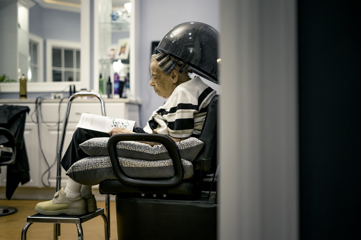A person sitting under a hair dryer in a salon, reading and relaxing.