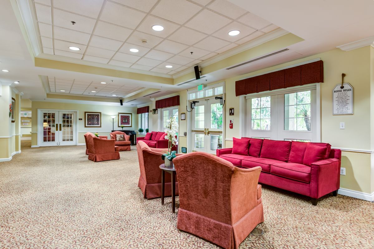 A spacious living room with comfortable seating, featuring red sofas and armchairs, carpeted flooring, large windows, and overhead lighting.