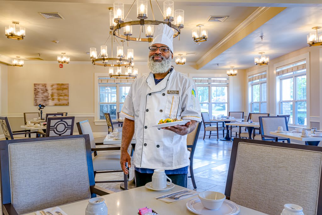 A chef smiling while holding a plate in an elegant dining room.