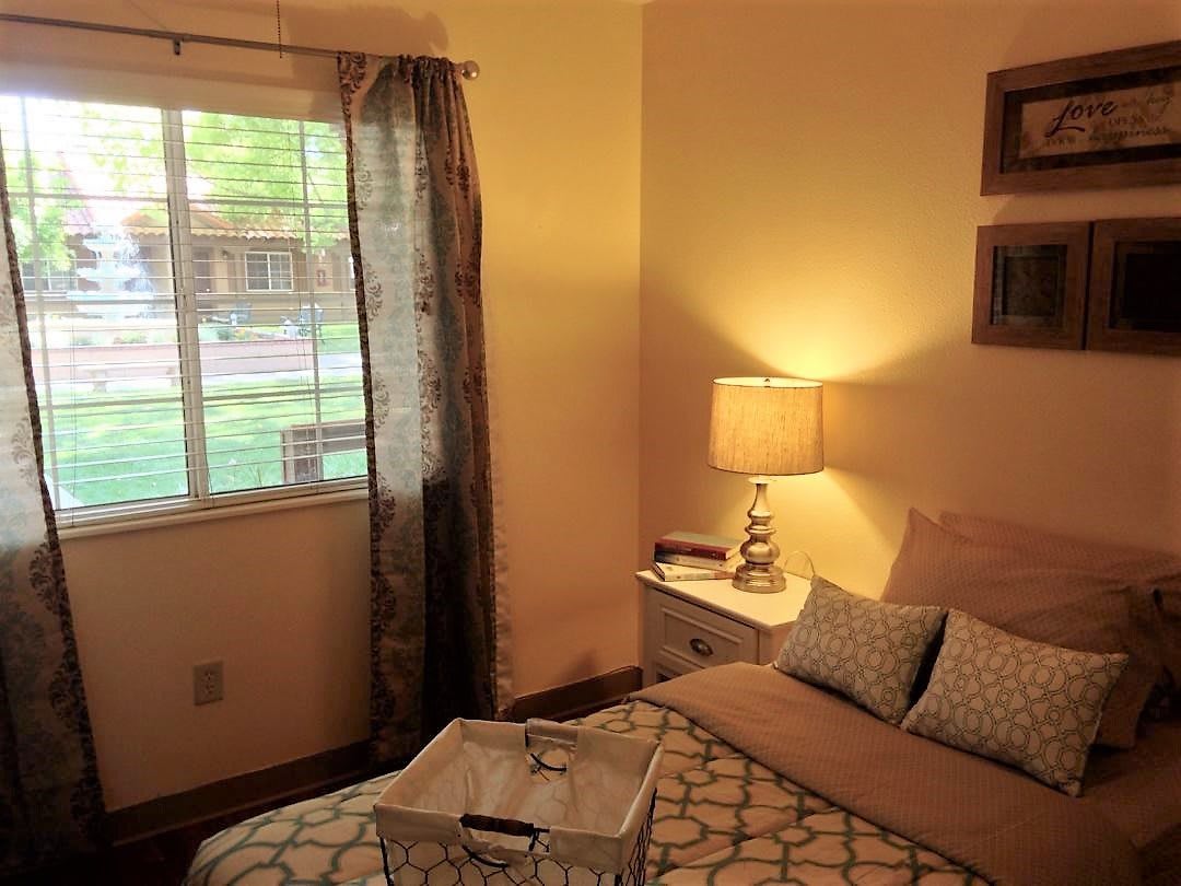 A cozy bedroom interior with a bed, nightstand, and table lamp by the window.